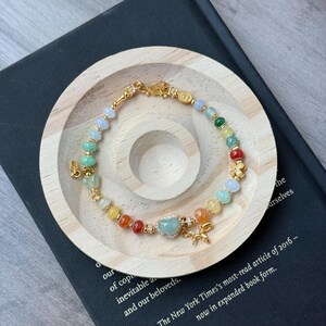 May include: A multi-colored beaded bracelet featuring round gemstone beads in shades of blue, green, yellow, orange, and red. The bracelet includes gold-toned metal accents, a central heart-shaped bead, and a small gold-toned balloon dog charm.