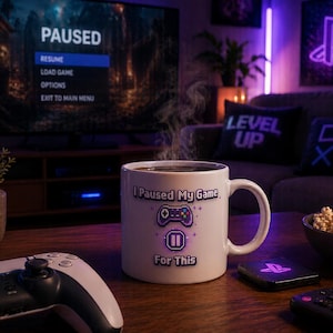 Könnte beinhalten: Eine weiße Kaffeetasse mit dem Text "I Paused My Game For This" und einem Controller-Motiv. Die Tasse steht auf einem Holztisch mit einem PlayStation 5-Controller, Popcorn und einem Fernseher, der ein pausiertes Spiel anzeigt.