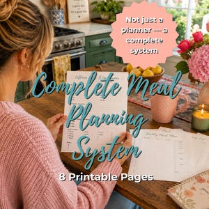 Op de afbeelding: Een vrouw in een roze trui houdt een maaltijdplanningssysteem vast met de tekst "Complete Meal Planning System" en "8 Printable Pages". Andere pagina's en een notitieboek liggen op de houten tafel. Een roze mok, kaars en bloemen zijn ook zichtbaar.
