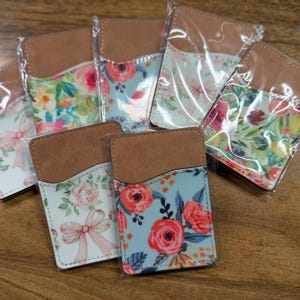 Puede incluir: Una colección de carteras para teléfono con estampado floral. Cada cartera presenta una parte superior de cuero marrón y un diseño floral colorido en la parte delantera. Los estampados florales incluyen rosas, lazos y otras flores en varios colores, como rosa, rojo y azul.