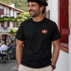 Puede incluir: Una camiseta negra de manga corta con un pequeño emblema de la bandera vasca en el pecho izquierdo, combinada con una boina negra y pantalones beige. La camiseta tiene cuello redondo y un corte estándar, mostrando el diseño de la bandera roja, verde y blanca.
