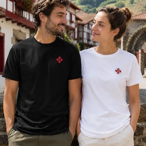 May include: A black short-sleeved t-shirt with a small red Basque cross emblem on the chest and a white short-sleeved t-shirt featuring a small red Basque cross emblem inside a circular border on the chest, both made of cotton fabric.