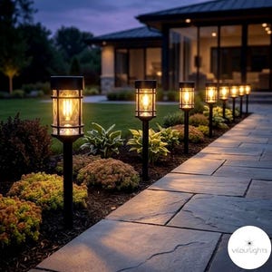 May include: A row of black outdoor pathway lights with a warm, inviting glow. Each light has a decorative cage design around the glass, illuminating a stone walkway lined with greenery. The lights are set against a backdrop of a house and a twilight sky.