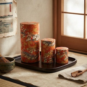May include: Three cylindrical canisters of varying heights featuring a vibrant orange background with a detailed floral and foliage pattern in white, gold, and dark tones. The canisters are arranged on a dark brown rectangular tray.