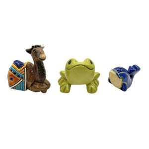 Pode incluir: Três figuras de cerâmica: um camelo marrom com uma sela colorida, um sapo verde e uma baleia azul. O camelo é detalhado com características esculpidas. O sapo tem um rosto sorridente. A baleia tem detalhes azuis e dourados.