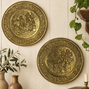 May include: Two circular brass wall plates featuring embossed scenes of figures gathered around tables. Each plate has a wide, decorative border with floral motifs. The metal has a polished, reflective gold-toned finish with intricate relief details.
