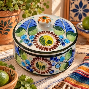 May include: A round ceramic lidded container featuring a hand-painted floral design with blue, green, yellow, and brown pigments on a white background. The lid has a central knob and is decorated with large blue leaves and a central flower motif.