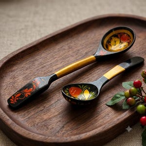 May include: Two wooden spoons with black and yellow painted handles and bowls. The larger spoon features a flat, rectangular handle end with orange floral patterns. The smaller spoon has a rounded bowl decorated with a red berry and yellow leaf motif.