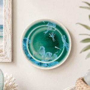 May include: A round ceramic plate featuring a deep emerald green glaze with a glossy finish. The surface is decorated with crystalline patterns in shades of light blue, forming star-like and fan-shaped clusters across the center and edges.