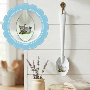 May include: A white metal decorative spoon featuring a painted illustration of a piglet resting in grass with a small blue bow. The spoon is suspended from a brass-colored hook, with a circular inset showing a detailed view of the piglet design.