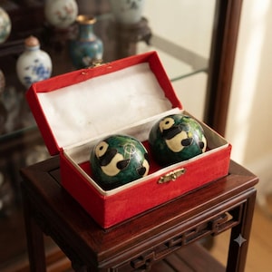 May include: Two spherical green meditation balls featuring black and white panda designs, housed in a rectangular red fabric-covered box with a white interior lining and a gold-toned metal latch closure mechanism on the front edge.