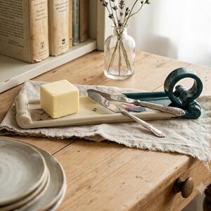 Puede incluir: Una mantequera crema con un cubo de mantequilla, dos cuchillos de mantequilla y un asa decorativa. La mantequera mide unos 20 cm de largo. Los cuchillos tienen mangos plateados ornamentados. Un pequeño jarrón de cristal con flores en el fondo.