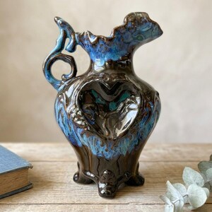 May include: A ceramic pitcher featuring a glossy dark brown and vibrant blue drip glaze finish. The vessel has a sculpted heart-shaped indentation on the front, a curved handle, a ruffled rim, and stands on three small feet with a decorative base.