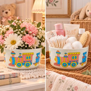 May include: A white ceramic bowl featuring a colourful graphic print of a pink and blue toy train with a small blue bear in the caboose. The bowl is shown filled with pink and white flowers in one view and bathroom essentials in the other.