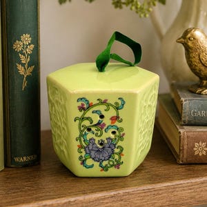 Peut inclure: Un récipient hexagonal en céramique vert clair avec un couvercle plat et une anse en ruban de velours vert. La face avant présente un motif floral coloré avec une fleur violette et trois petits trous circulaires pour la ventilation.