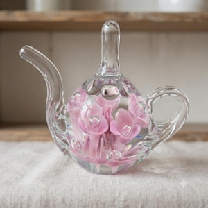 May include: A clear glass teapot-shaped paperweight featuring internal pink floral-like shapes and small air bubbles suspended within the glass. The object has a curved spout, a central vertical handle, and a rounded body with a smooth finish.