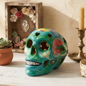 May include: A teal ceramic skull sculpture featuring hand-painted floral patterns in red, yellow, and white. The skull has cut-out eye sockets, a nose cavity, and a mouth with black and white teeth detailing, finished with a glossy glaze.