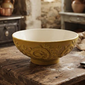 May include: A round ceramic mixing bowl with a mustard yellow exterior featuring a raised, embossed scrollwork pattern. The interior of the bowl is a smooth, solid off-white color. The bowl has a wide, open top and a sturdy, circular base.