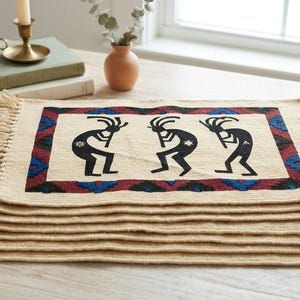 Può includere: Una pila di tovagliette rettangolari intrecciate di colore beige chiaro con bordi sfrangiati. Ogni tovaglietta presenta un motivo centrale con tre figure nere di Kokopelli e un bordo decorativo nei toni del rosso scuro, blu e nero.
