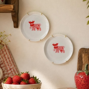 May include: Two white ceramic plates mounted on a wall, each featuring a red graphic of a cow with a checkered pattern holding a small flower. The plates are circular with smooth edges and are held in place by simple gold-toned metal wire clips.