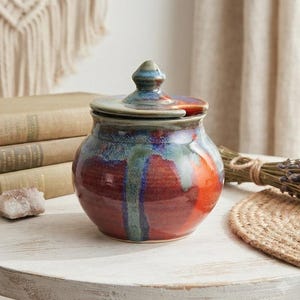 May include: A round ceramic jar with a lid featuring a glossy glaze in shades of deep red, burnt orange, and blue-green. The jar has a wide, bulbous body and a small, pointed knob on the centre of the lid, showcasing a smooth, reflective finish.
