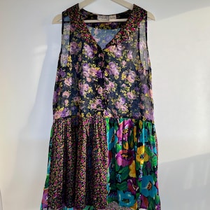 May include: A sleeveless dress featuring a button-down front and a V-neckline. The bodice has a purple and yellow floral pattern on a black background. The skirt is divided into two panels with small floral and large vibrant floral prints.