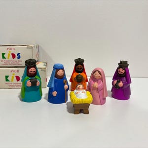 May include: A set of Avon Kids My First Christmas Story Nativity Collection figurines including five figures and a baby in a manger. The figures are dressed in colorful robes of teal, blue, orange, pink, and purple, with two original cardboard boxes.