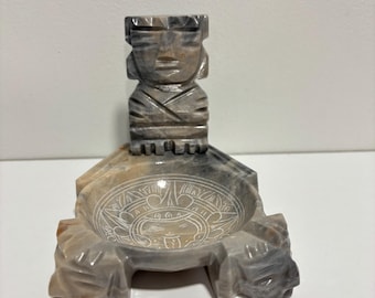 May include: A gray and tan stone bowl featuring a carved figure standing at the back and an etched circular design at the center of the basin. The bowl is supported by three carved stone feet, showcasing natural marbling and smooth textures.