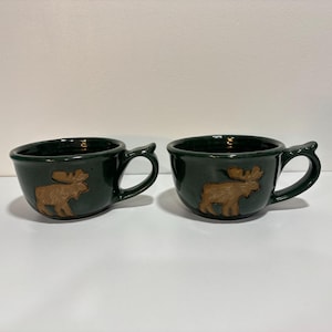 May include: Two dark green ceramic mugs, each featuring a raised, light brown relief of a moose on the front. The mugs have a wide, rounded bowl shape and a curved handle on the right side, with a smooth, glossy finish throughout.
