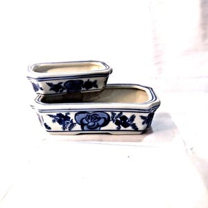 May include: Two rectangular ceramic planters with rounded corners, featuring a white base and a blue floral pattern. The larger planter sits at the bottom, while the smaller planter is stacked on top, both displaying hand-painted blue roses.