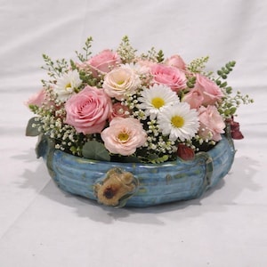 Peut inclure: Un arrangement floral composé de roses rose clair, de marguerites blanches et de petites fleurs blanches dans un bol en céramique bleu. Le bol présente une surface texturée avec une petite figurine de tortue brune sculptée sur le bord avant.