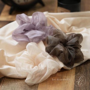 May include: Three sheer fabric hair scrunchies in lavender, charcoal gray, and cream colors. Each scrunchie features a gathered, ruffled texture and a lightweight, semi-transparent appearance. The text Lilt & Charm Co. is visible in the corner.