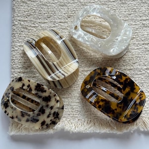 May include: Four oval hair clips with a smooth, glossy finish. One clip is white with a marbled pattern, one is beige with dark brown vertical stripes, one is cream with dark brown spots, and one is dark brown with a classic tortoiseshell pattern.