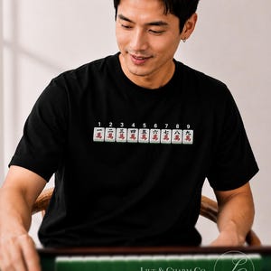 May include: A black short-sleeved t-shirt featuring a graphic print of nine mahjong tiles arranged in a horizontal row. Each tile displays a number from one to nine in black above a red Chinese character. The shirt has a crew neckline and short sleeves.