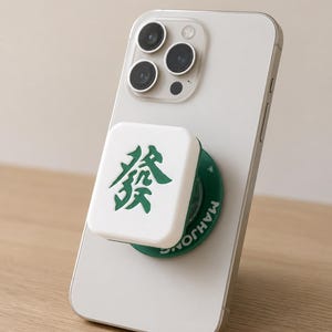May include: A white smartphone with a square, white phone grip attached to the back. The grip features a green Chinese character for 'Fa' on its face and a green circular base with the word 'MAHJONG' printed in white letters along the side.