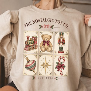 May include: A beige crewneck sweatshirt featuring a graphic print with the text THE NOSTALGIC TOY CO. and EST. 1994. The design includes six rectangular panels displaying a toy train, a teddy bear, a nutcracker, a spinning top, a star, and a candy cane.