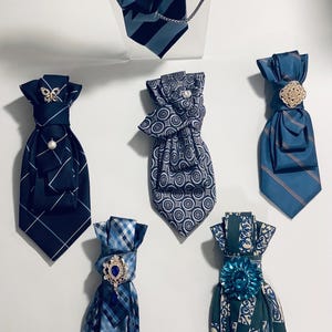 May include: A collection of six decorative neck cravats in various blue and green shades. Each cravat features distinct patterns including stripes, geometric circles, plaid, and floral motifs, accented with ornate crystal and metal brooches.