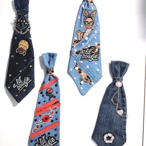 May include: Four denim neckties featuring various embellishments. One dark blue tie has gold studs and a chain. One light blue tie features a leopard print head graphic, pearls, and the text a lil bougie. One tie has red zippers, roses, and text. One tie has a chain and pearl.