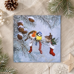 May include: A square paper napkin featuring a winter scene with three birds perched on pine branches. A robin, a blue tit, and a wren are depicted alongside three hanging Christmas stockings. The background shows falling snow and pine cones.