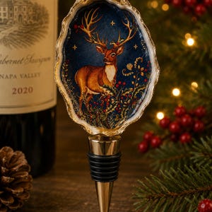 May include: A bottle stopper featuring a decorative oyster shell top with a painted scene of a brown deer standing among red holly berries against a dark blue starry background. The shell has a gold-painted rim and a metallic base with black rings.