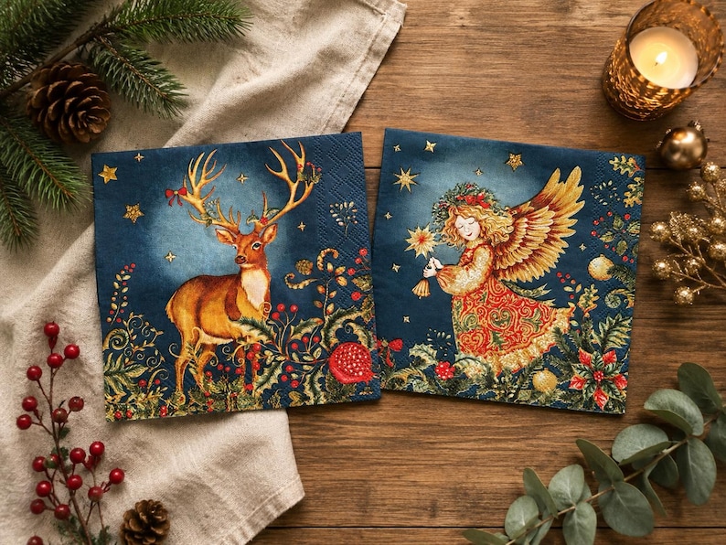 May include: Two square paper napkins featuring festive holiday illustrations on a dark blue background. One napkin displays a brown deer with large antlers, while the other shows an angel with golden wings holding a star, both surrounded by greenery.