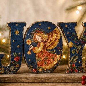May include: A decorative wooden sign spelling the word JOY in dark blue. The letter O is an oval featuring an illustration of an angel with golden wings and a patterned dress, surrounded by stars, holly leaves, and red berries on all letters.