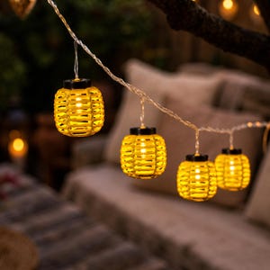 May include: A string of four yellow lantern-shaped lights hanging from a thin wire. Each lantern features a woven, cage-like exterior with a black top cap and a warm, glowing light source visible inside the center of the structure.
