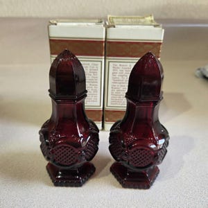 May include: A pair of deep red glass salt and pepper shakers featuring a faceted, hexagonal base and a textured, diamond-patterned body. Each shaker has a pointed, multi-faceted lid and is positioned in front of its original white cardboard box.