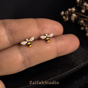 Puede incluir: Un par de pendientes de botón con forma de abeja dorada, con alas de cristales transparentes y cuerpo a rayas amarillas y negras. El texto ZalfahStudio es visible en la parte inferior. Estos pequeños pendientes tienen un acabado pulido.