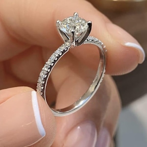 May include: A close-up of a diamond engagement ring. The ring features a large, round-cut diamond set in a four-prong setting. The band is made of white metal and is encrusted with smaller diamonds. The ring is held up by a person.