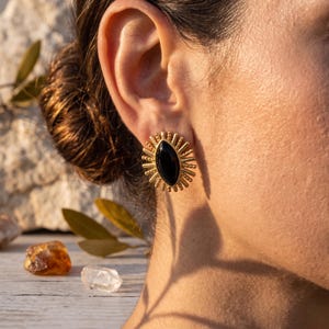 Peut inclure: Boucles d'oreilles dorées avec une pierre noire ovale au centre. Les boucles d'oreilles ont un motif de rayons de soleil autour de la pierre. Les boucles d'oreilles sont sur l'oreille d'une personne.