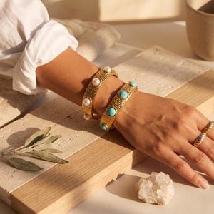 Peut inclure: Deux bracelets manchettes en métal doré avec une surface texturée et martelée. L'un des bracelets est orné de perles blanches rondes, tandis que l'autre est serti de pierres turquoise rondes. Une bague en métal doré torsadé est portée.