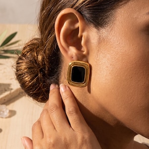 May include: Gold-coloured square earrings with a black centre. The earrings have a polished finish and a classic design. The image shows a close-up of the earrings on an ear, highlighting the jewellery's details and elegant style.