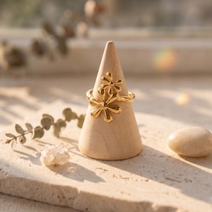 Peut inclure: Une bague dorée ornée de deux formes de fleurs stylisées superposées aux pétales arrondis. La bague est présentée sur un support conique en bois clair et lisse, mettant en valeur la finition métallique polie et le design organique et superposé.